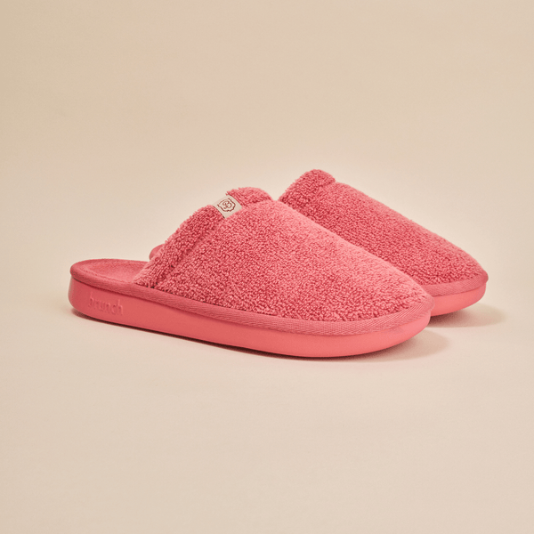Three-quarter view of Essential Slipper in coral pink terry showing plush upper, open back and cushioned sole on beige
