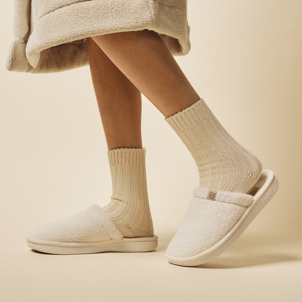 Side view of Essential Spring slipper with cream terry upper and cushioned sole worn with ribbed socks on cream background