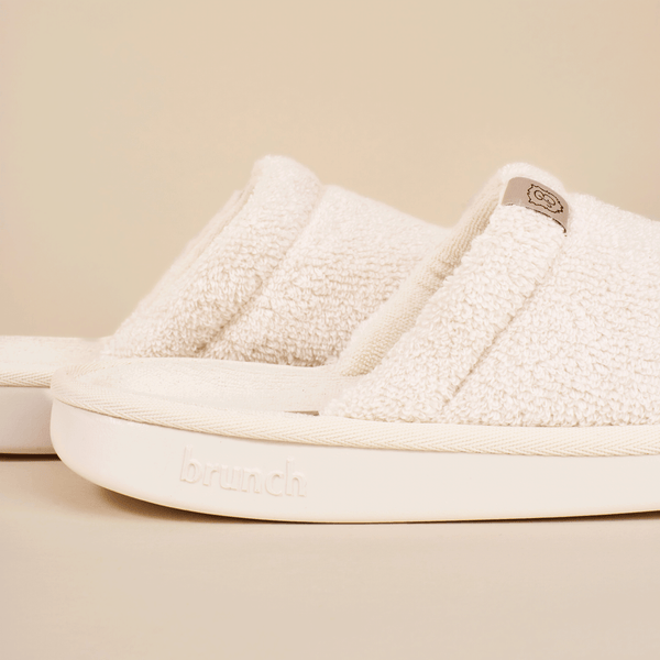 Close-up side view of cream Essential slipper showing terry-cloth upper and embossed brunch logo on the sole on a beige background