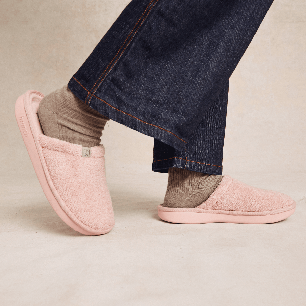 Side view of pink plush slip-on slipper showing fuzzy terry upper and molded sole on model feet with denim hem, neutral background
