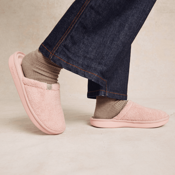 Side view of pink plush slip-on slipper showing fuzzy terry upper and molded sole on model feet with denim hem, neutral background