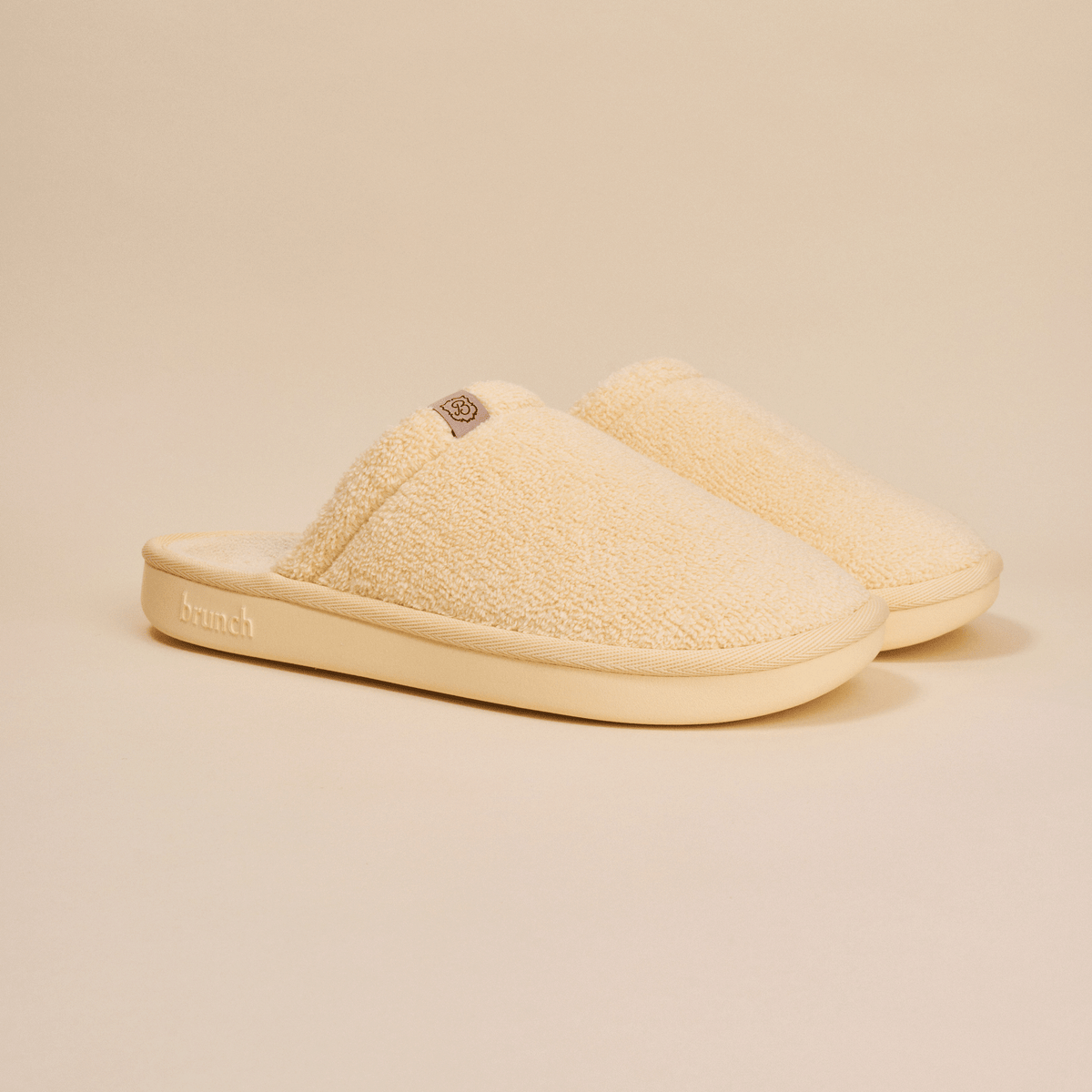 Three-quarter view of Essential slipper showing cream sherpa upper, slip-on style, and cushioned sole with embossed Brunch logo on a neutral background