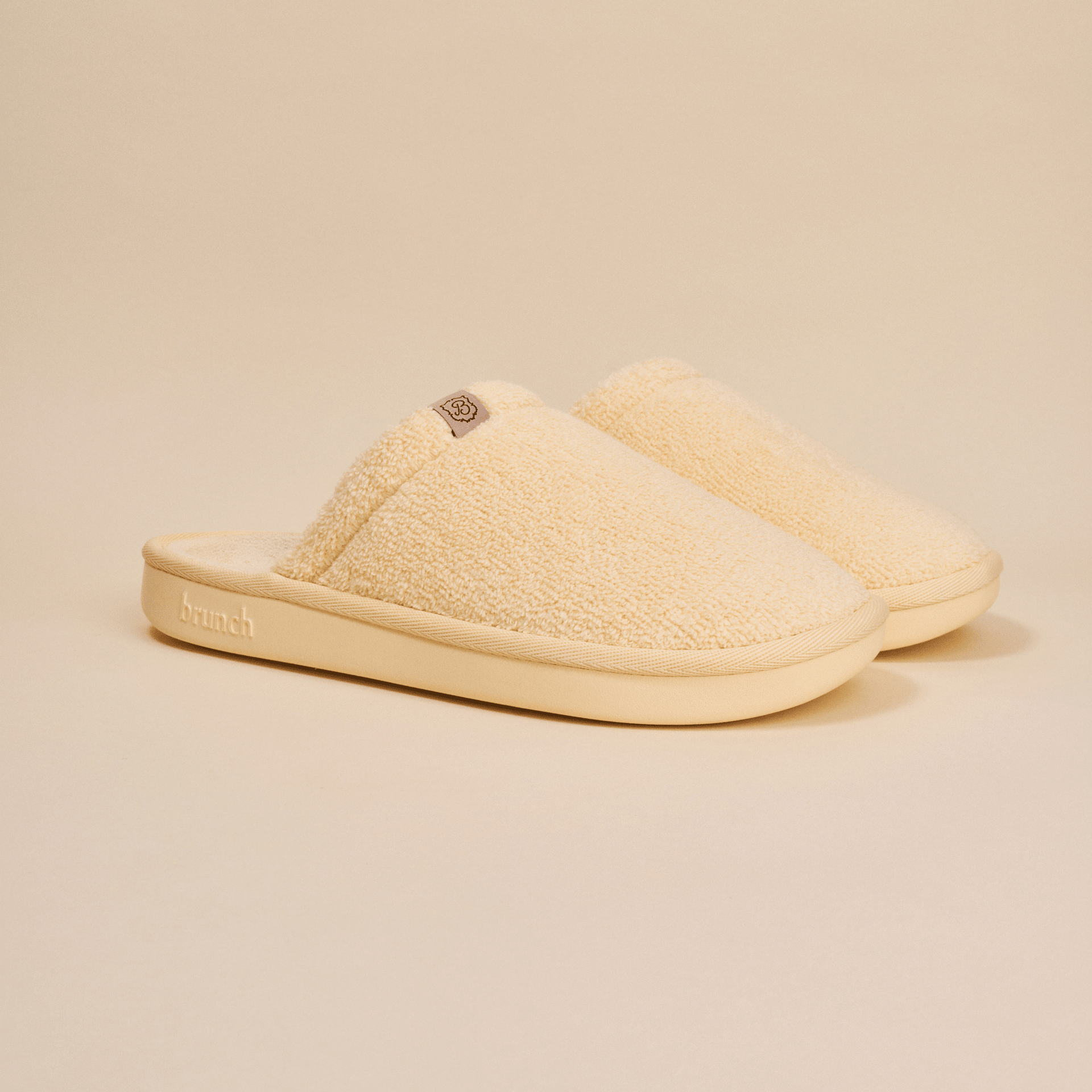 Three-quarter view of Essential slipper showing cream sherpa upper, slip-on style, and cushioned sole with embossed Brunch logo on a neutral background