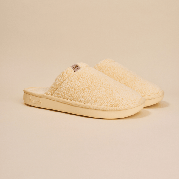 Three-quarter view of Essential slipper showing cream sherpa upper, slip-on style, and cushioned sole with embossed Brunch logo on a neutral background