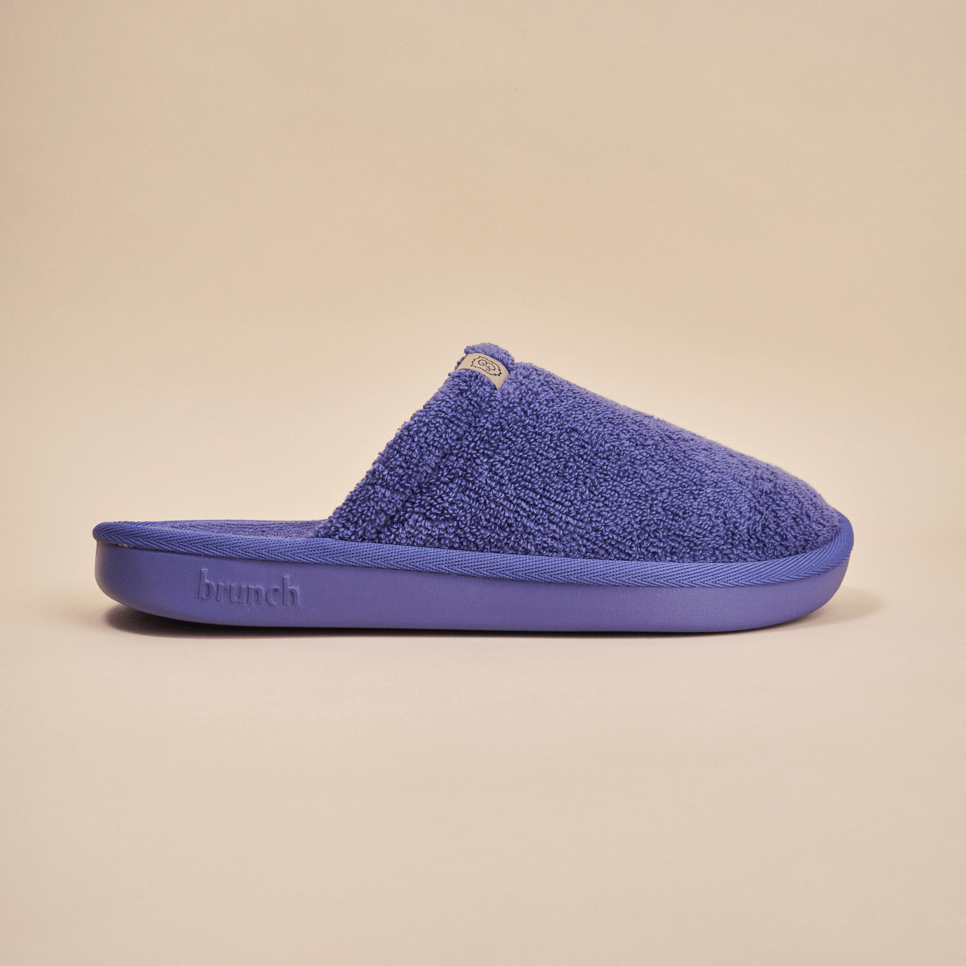 Side view of Essential slipper showing purple terry upper and molded sole embossed with brunch logo on beige background