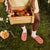 Model holding a wooden crate of strawberries wearing coral pink fuzzy Brunch slippers and white socks on a grassy, flower-strewn backdrop