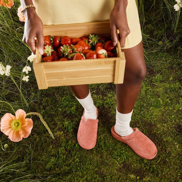 Model holding a wooden crate of strawberries wearing coral pink fuzzy Brunch slippers and white socks on a grassy, flower-strewn backdrop