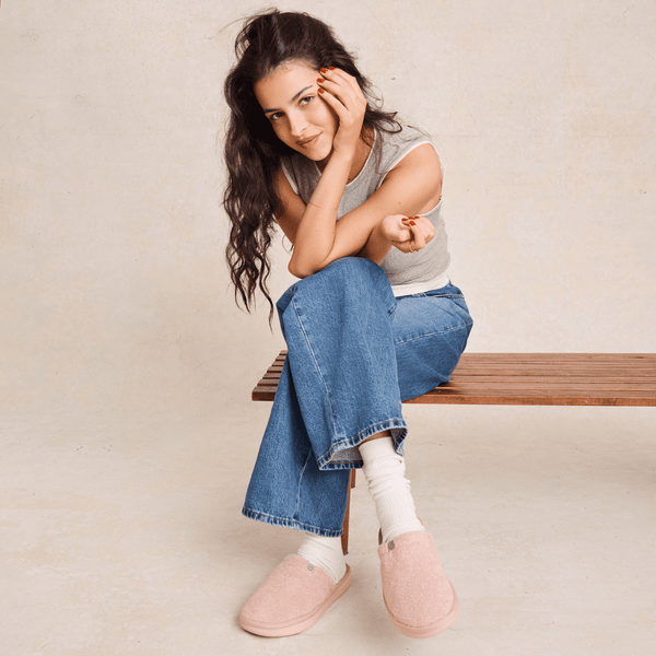 Three-quarter view of Essential Slipper in blush pink showing terry upper and cushioned sole worn by model on a bench