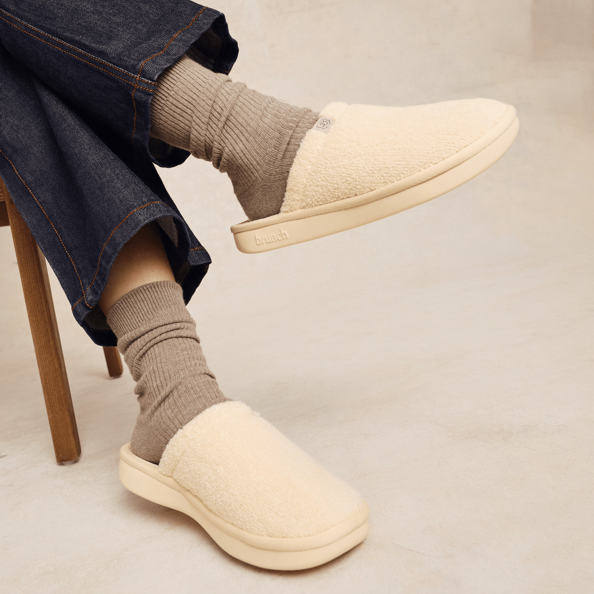 Side view of Essential slipper in cream showcasing sherpa upper and cushioned sole with embossed brunch logo, worn with socks