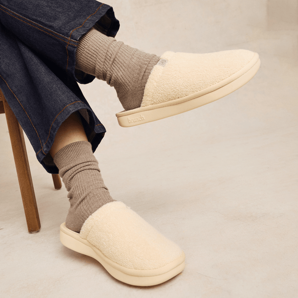 Side view of Essential slipper in cream showcasing sherpa upper and cushioned sole with embossed brunch logo, worn with socks