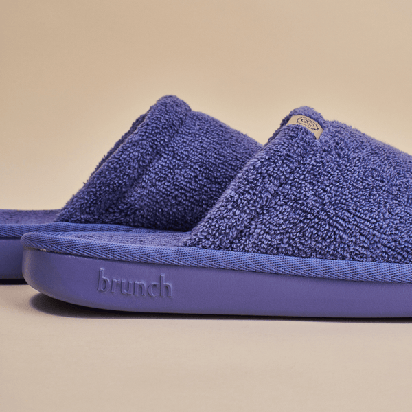 Close-up side view of Essential slipper in purple showing terry cloth upper and molded sole embossed with brunch on beige background