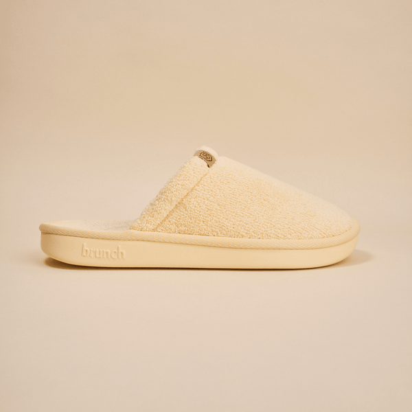Side view of Essential Slipper showing cream sherpa upper and cushioned sole with embossed brunch logo on neutral background