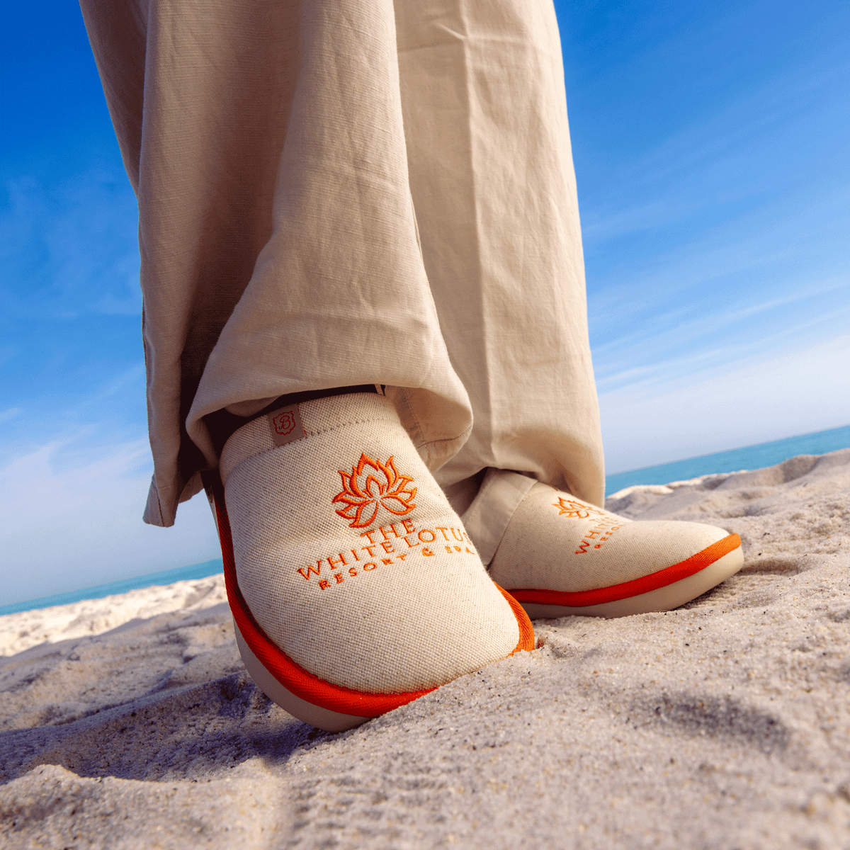 Low-angle front view of Essential The White Lotus slipper in beige showing orange embroidered lotus logo and EVA outsole on sand