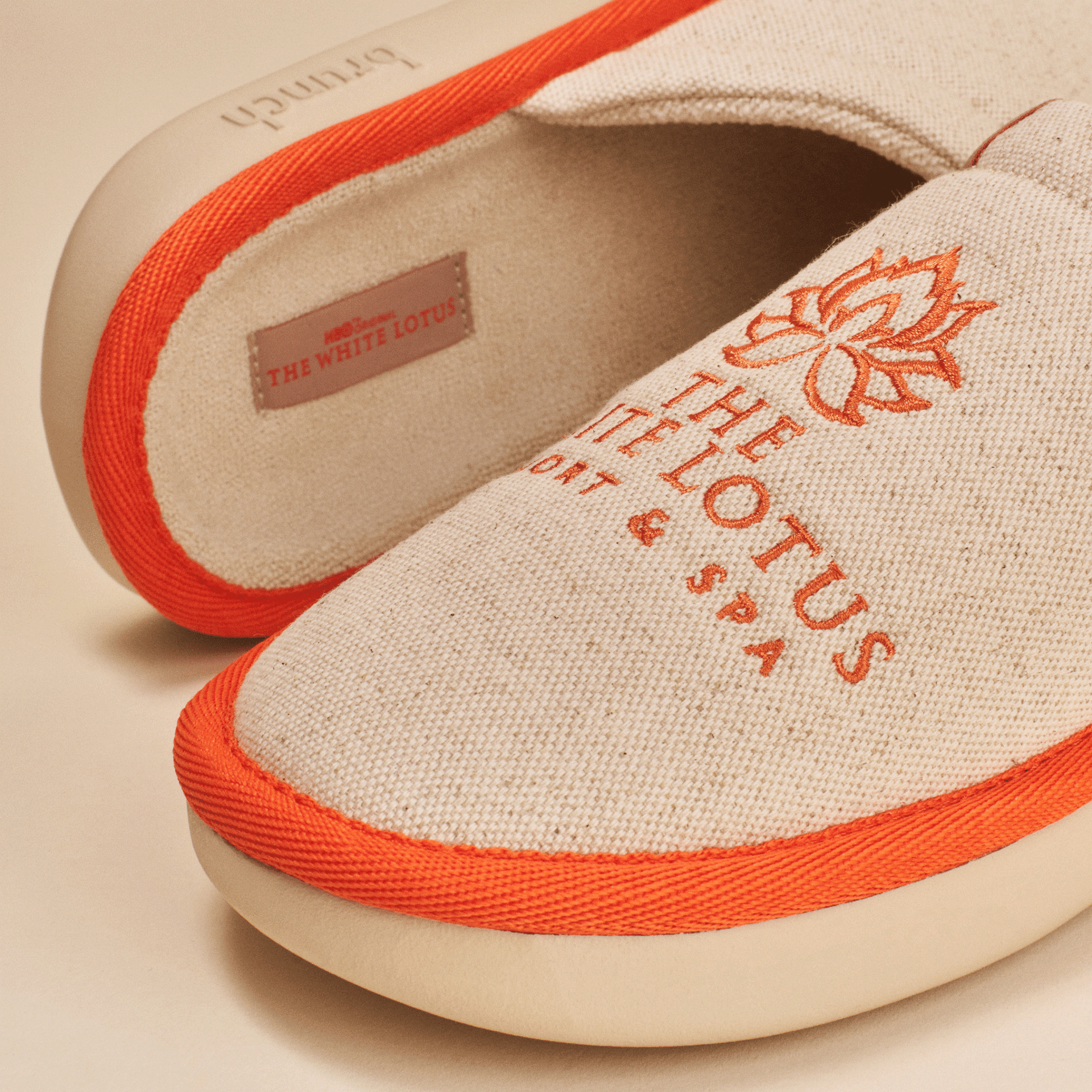Close-up of The White Lotus slipper showing beige linen upper with orange embroidered lotus logo, orange piping and cushioned EVA sole