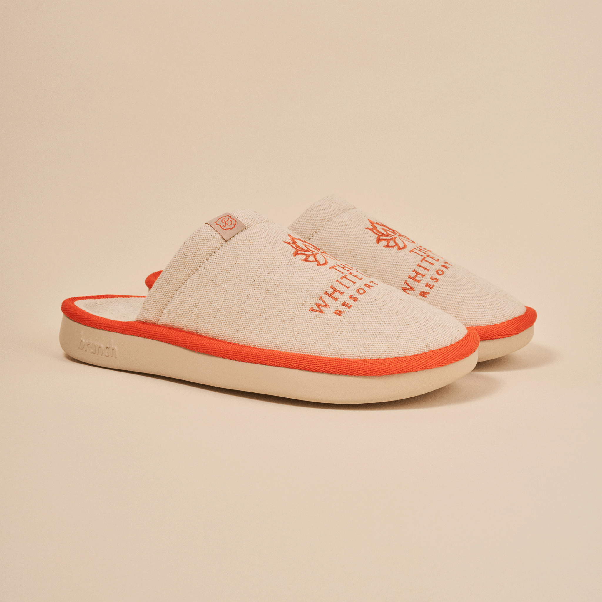 Angled view of The White Lotus slipper in beige with embroidered THE WHITE LOTUS RESORT, linen upper and orange trim