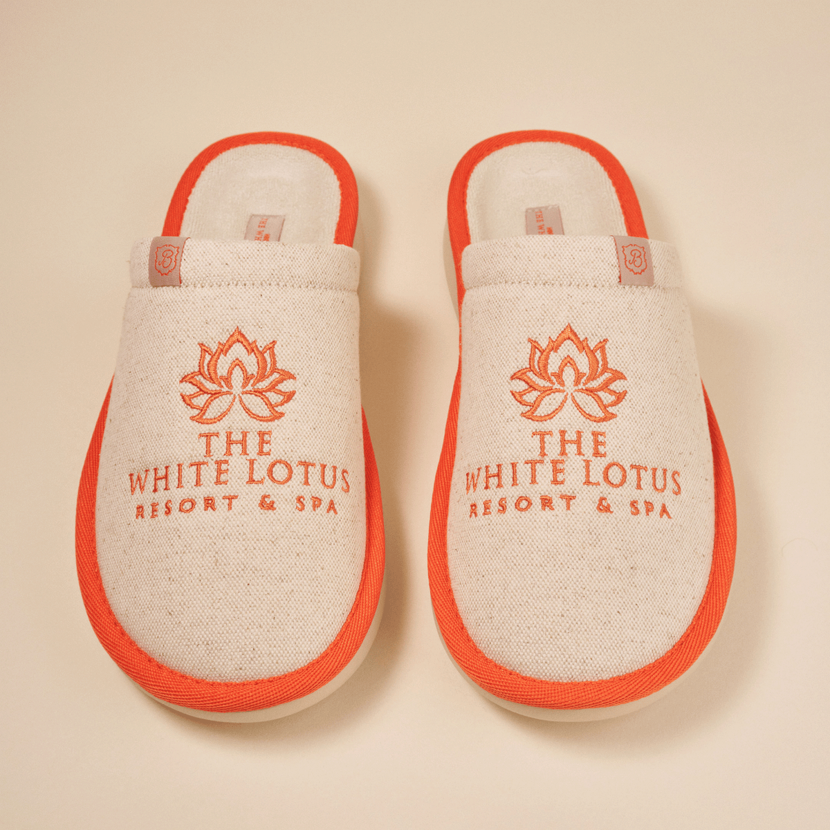 Top-down view of Essential The White Lotus linen slippers in beige with orange trim, embroidered THE WHITE LOTUS RESORT & SPA logo