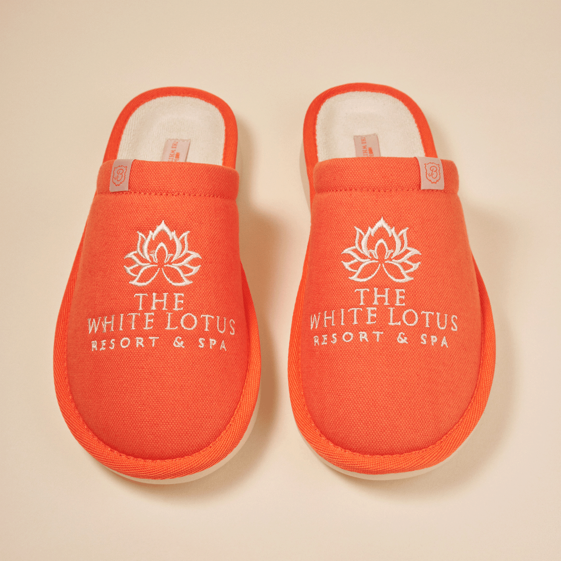 Top view of pair of orange White Lotus Essential slippers showing white embroidered 'THE WHITE LOTUS RESORT & SPA' logo and terry lining on a beige background