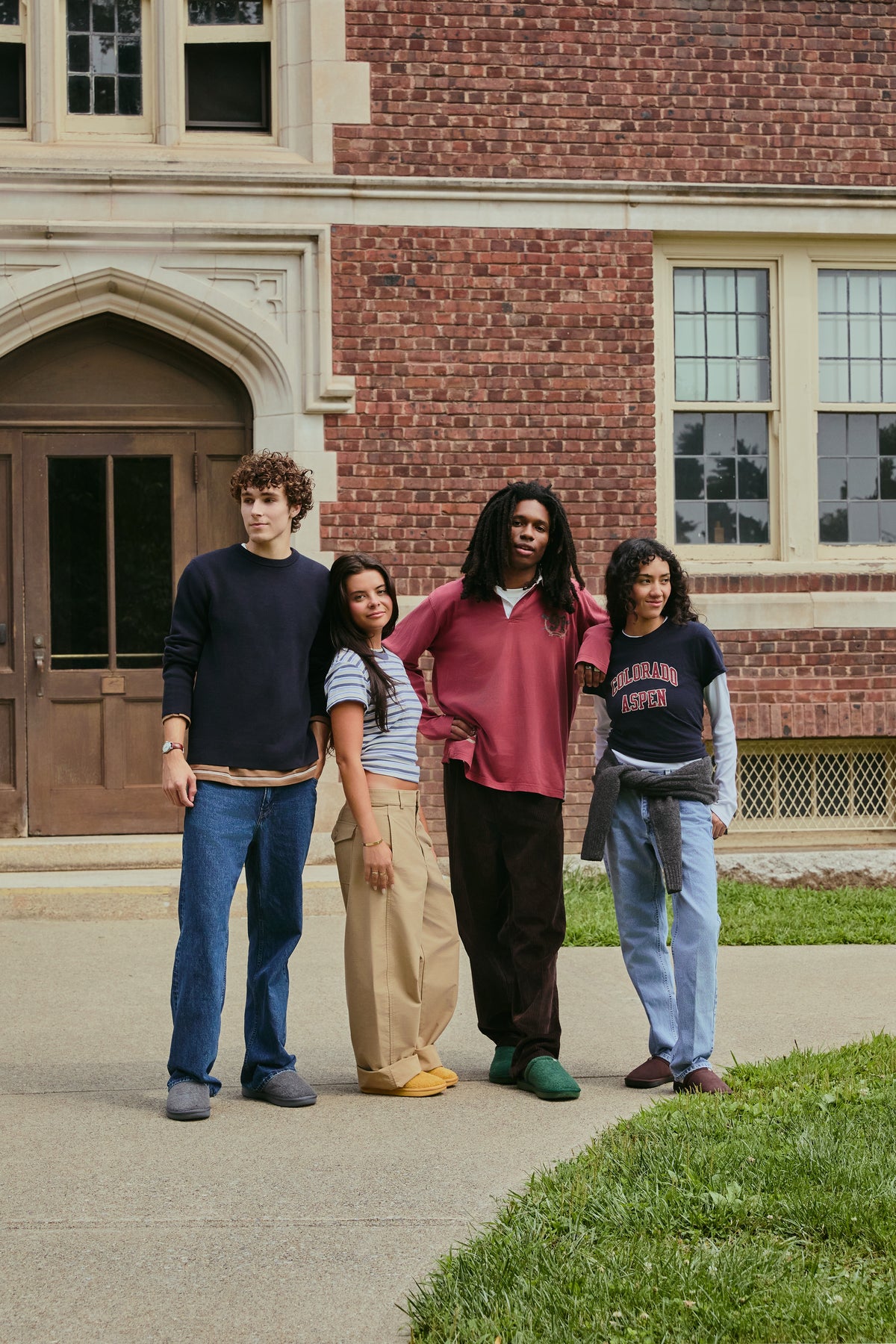 Four college students standing on a brick campus quad wearing colorful slippers and casual fall outfits