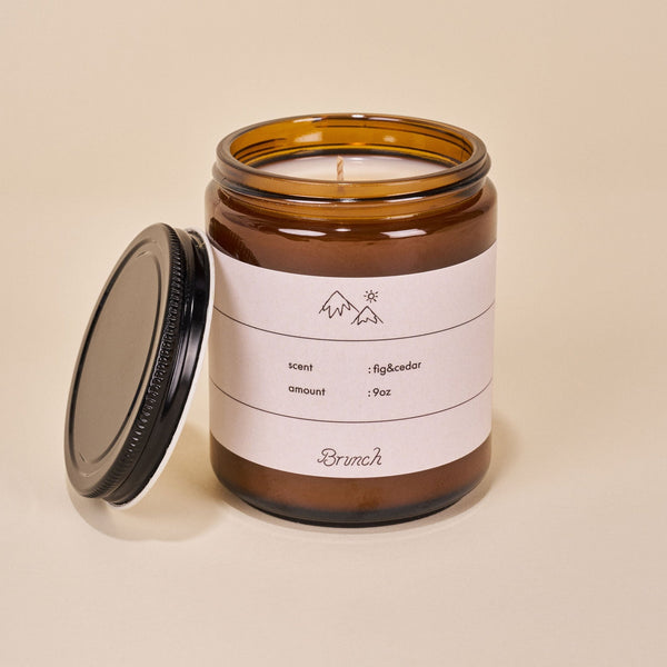 Three-quarter view of Holiday Candle in amber glass jar showcasing label fig&cedar 9oz with lid beside on beige background
