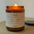Close-up front view of Holiday Candle (Cedar) in amber glass with lit flame and label reading scent fig and cedar, amount 9oz on wooden table