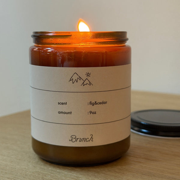 Close-up front view of Holiday Candle (Cedar) in amber glass with lit flame and label reading scent fig and cedar, amount 9oz on wooden table