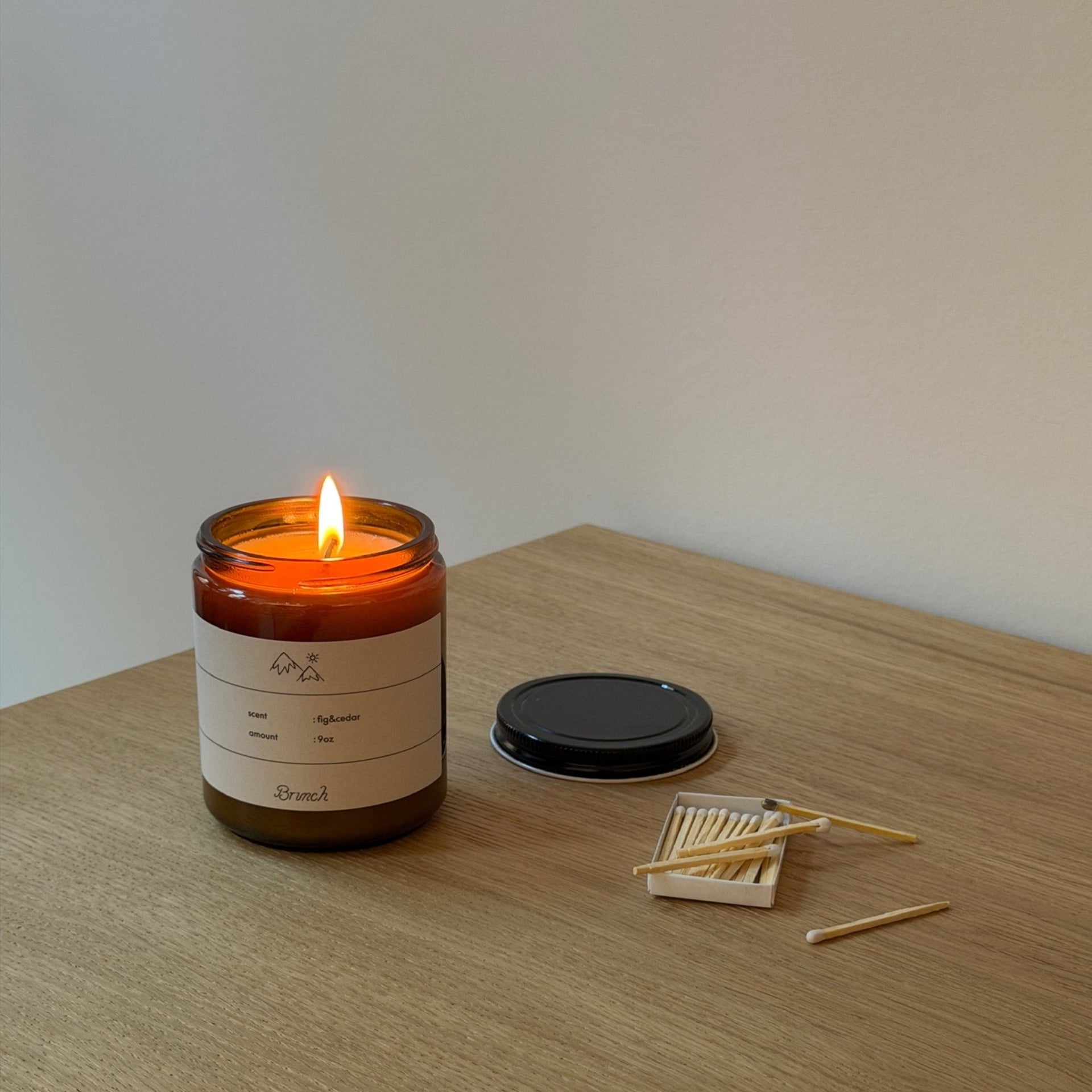 Three-quarter view of Brunch Fig and Cedar holiday candle in amber jar with lit flame, lid and matchbox on a wood table