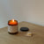 Three-quarter view of Brunch Fig and Cedar holiday candle in amber jar with lit flame, lid and matchbox on a wood table