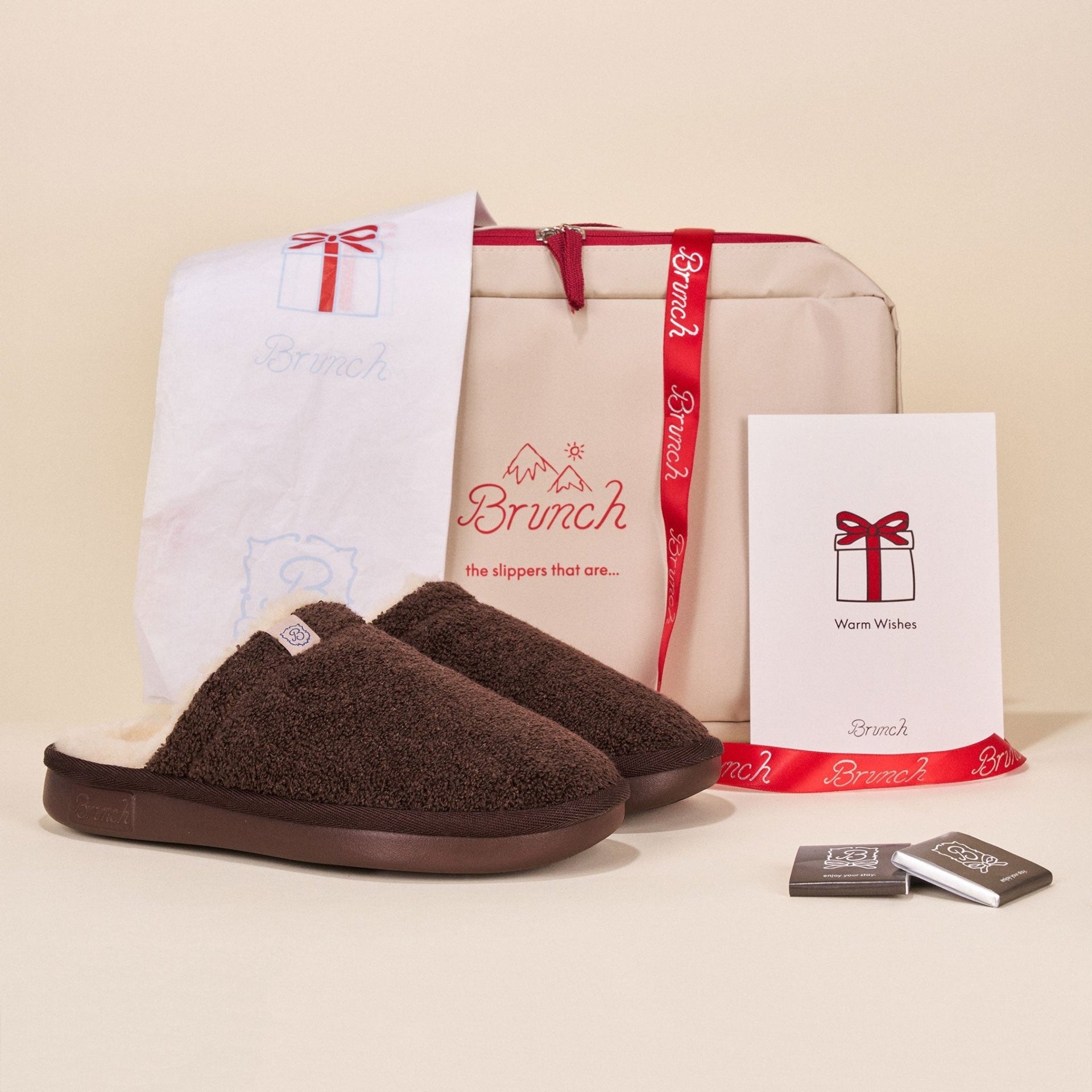 Front view of Holiday Packaging set with brown fuzzy slippers and Brunch gift bag with red ribbon on cream background