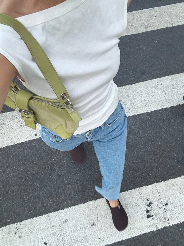 Person walking across crosswalk wearing white tank top, light blue jeans, dark slip-on shoes and small green shoulder bag.