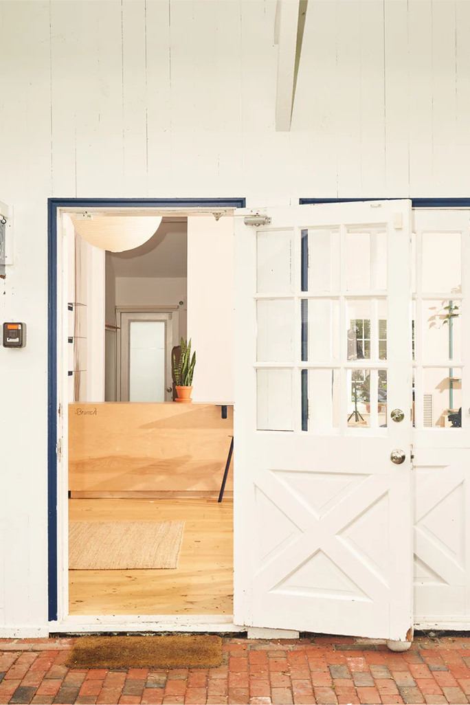 Open white French door to a bright entry with a wooden reception desk labeled 'Branch', a potted snake plant, rug, and brick porch.