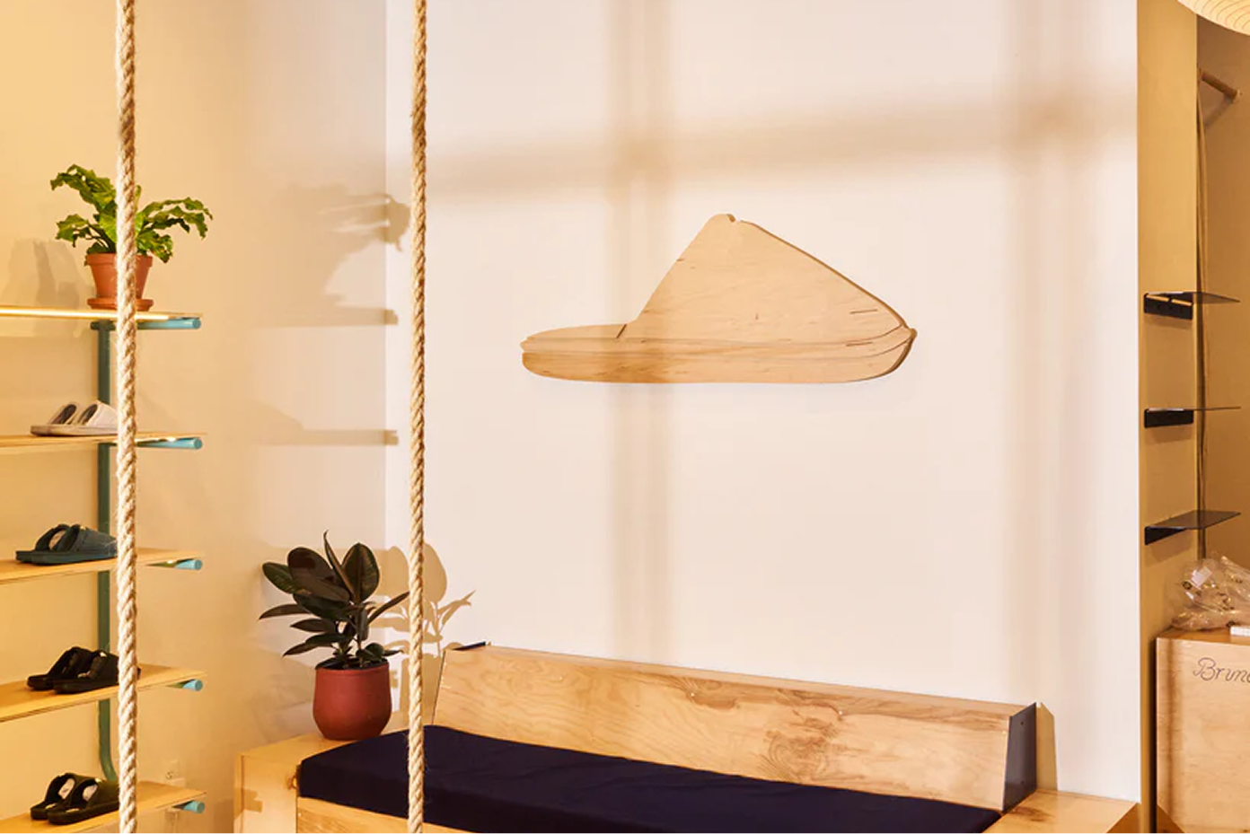Retail interior: wooden bench with navy cushion, hanging rope swings, potted plant, shoe shelves and minimal wood wall art.