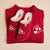 Top-down view of folded crimson Parke mockneck fleece with matching slippers embroidered 'parke' and 'brunch' with three hearts on a pale beige background