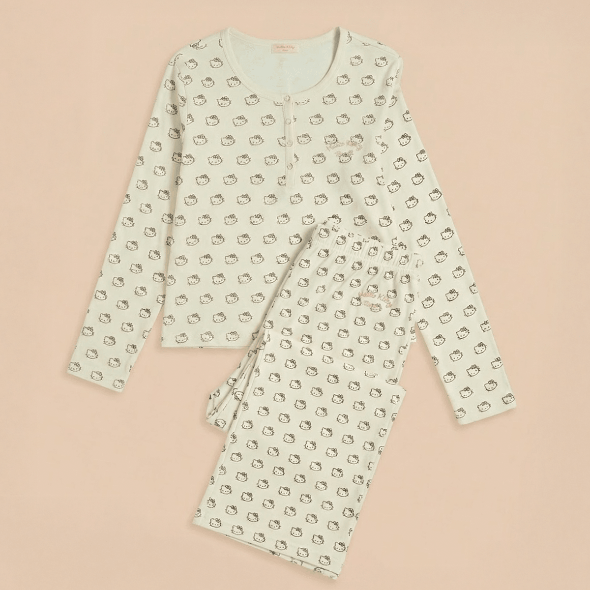 Flat lay of cream Hello Kitty cotton pajamas, long-sleeve button top and elastic-waist pants on pale pink background