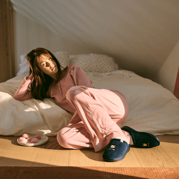 Model lounging on bed in a pink Hello Kitty pajama set with navy embroidered Hello Kitty slippers in warm morning bedroom light