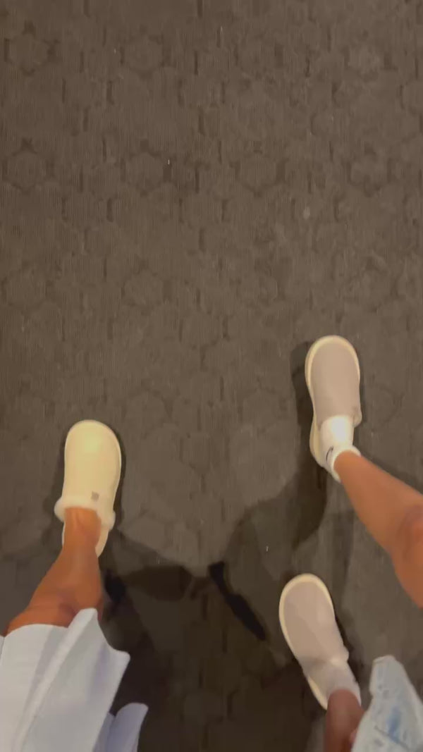 Overhead view of three people's feet wearing beige slip-on shoes and white socks on dark hex-patterned carpet.