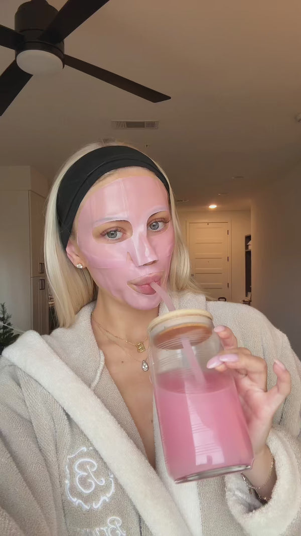 Person wearing pink sheet face mask and black headband, in a bathrobe, sipping a pink drink from a glass jar through a straw.