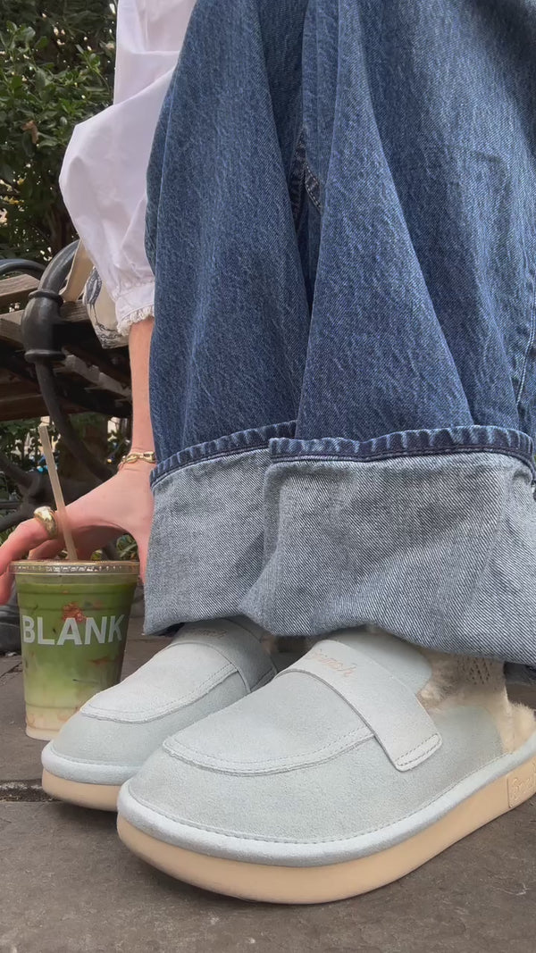 Cuffed denim, light-blue shearling-lined slip-on shoes, and a hand reaching for a green iced drink in a cup labeled BLANK.