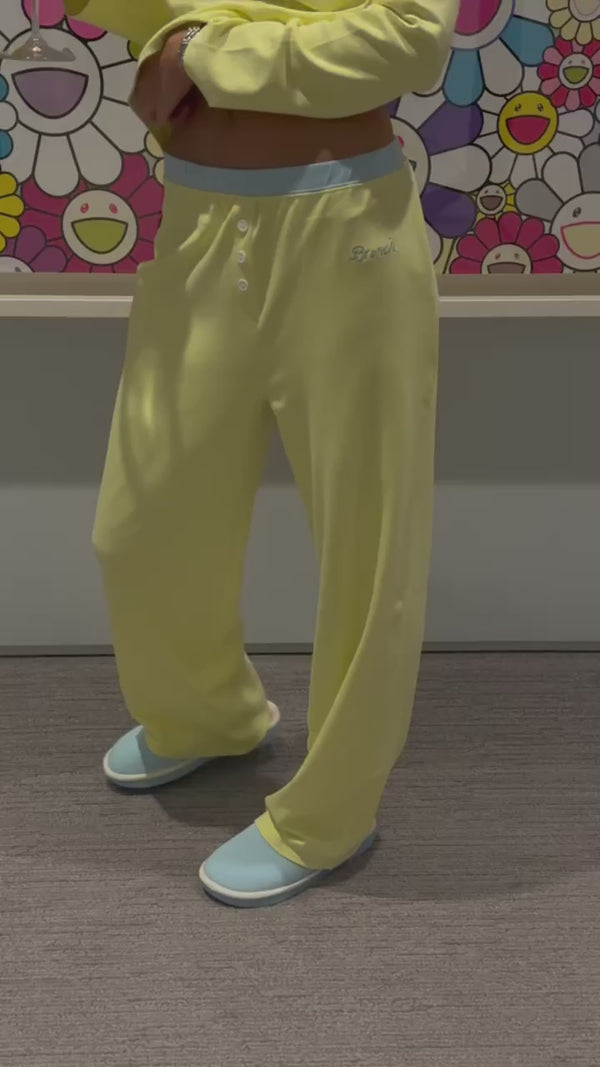Person wearing pale yellow wide-leg lounge pants and light blue slippers, cropped midriff visible, standing before colorful smiling-flower mural.