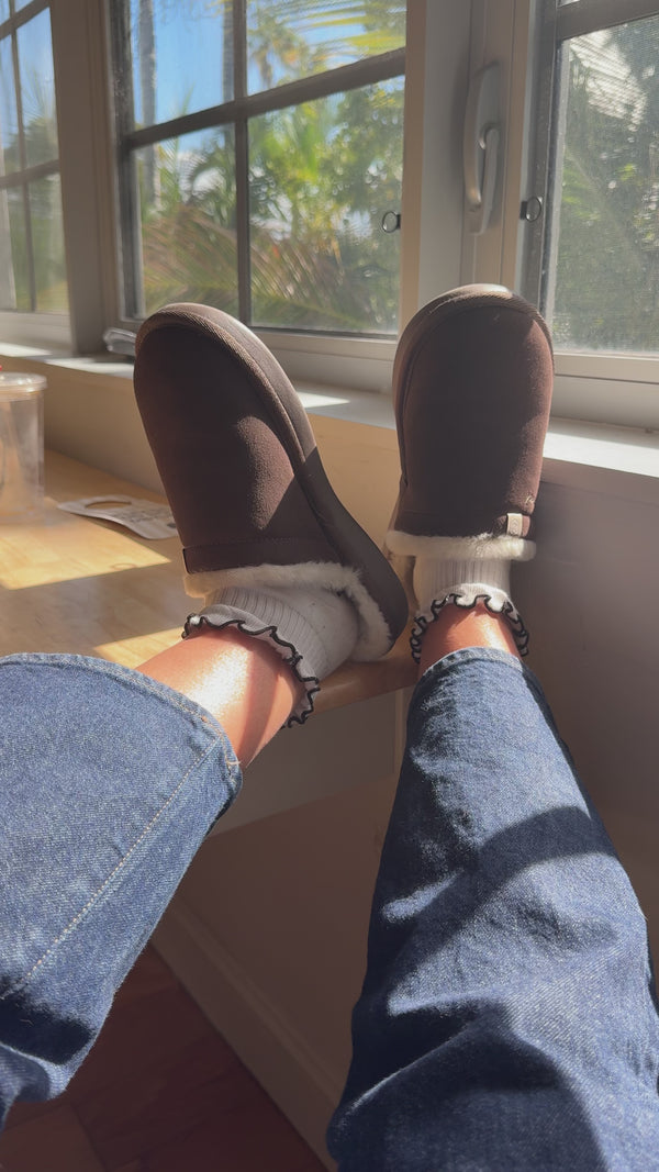 Legs propped on a sunlit windowsill wearing brown fleece-lined slip-on slippers, ruffled white ankle socks and blue jeans