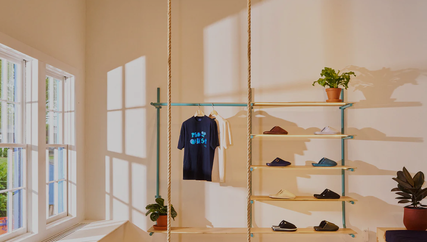 Sunlit retail display with rope-hung rack, navy T-shirt reading PLAY HOUSE, slippers on shelves and potted plants.