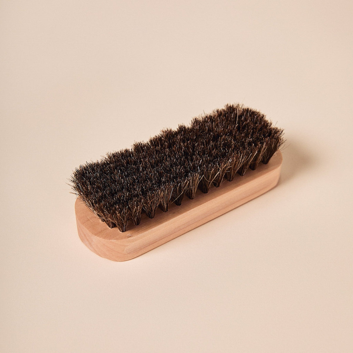 Angled top view of suede brush showing dense dark horsehair bristles and light wood handle on a pale beige background