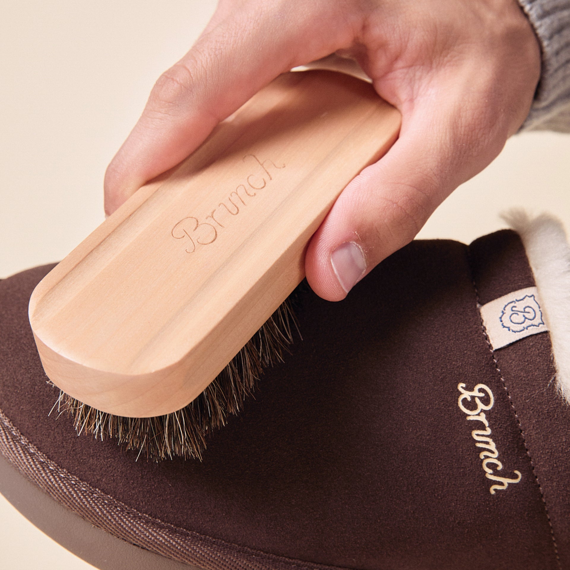 Hand holding a wooden shoe brush engraved Brunch, brushing a brown slipper embroidered Brunch.