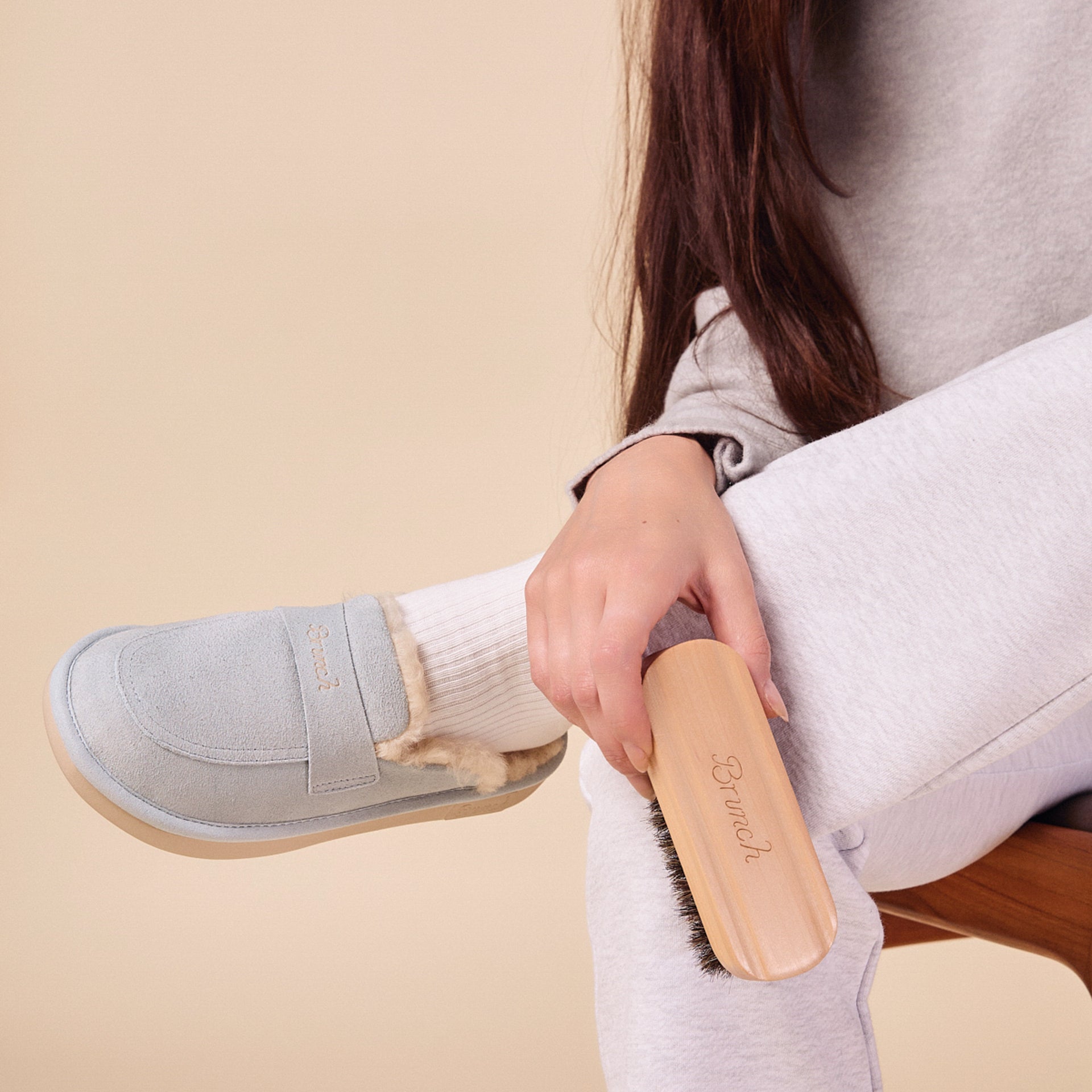 Hand holding wooden Brunch-branded shoe brush brushing a gray shearling-lined slipper