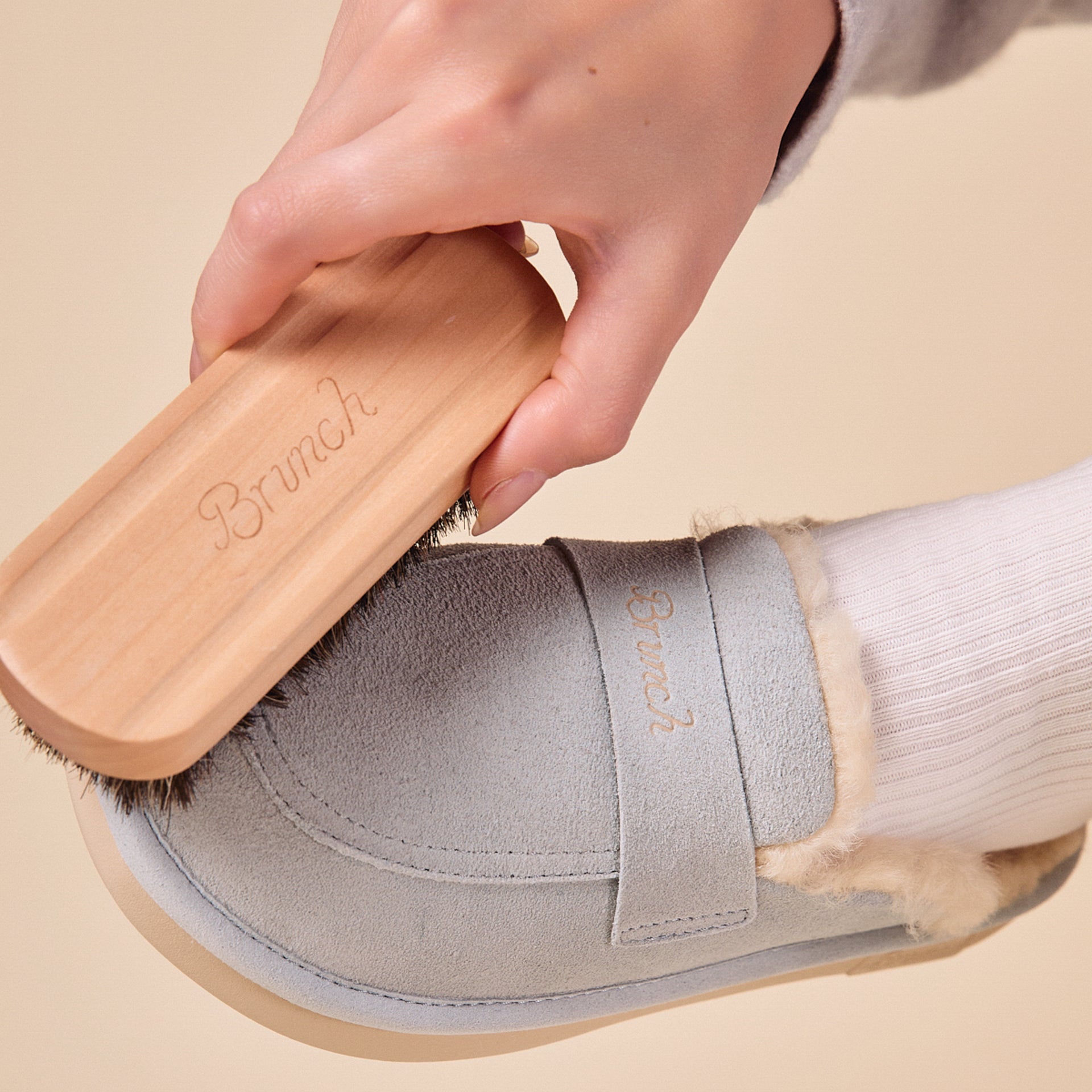 Hand holding wooden brush engraved 'Brunch' brushing a gray suede slipper with 'Brunch' on the strap.