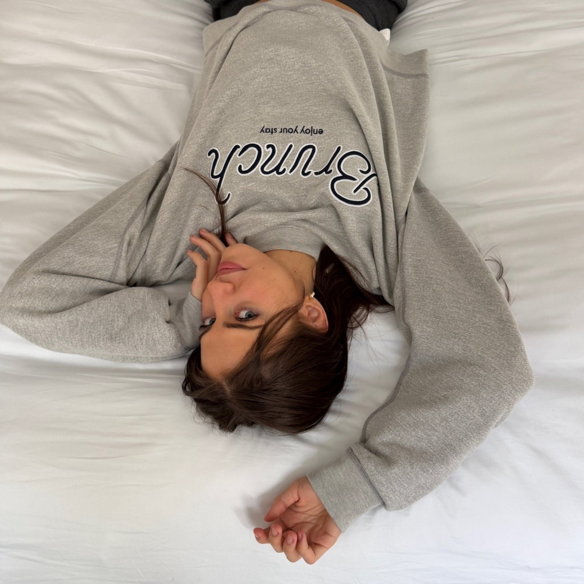 Woman lying on white sheets wearing a cropped heather grey varsity crewneck in French terry with Brunch chest lettering