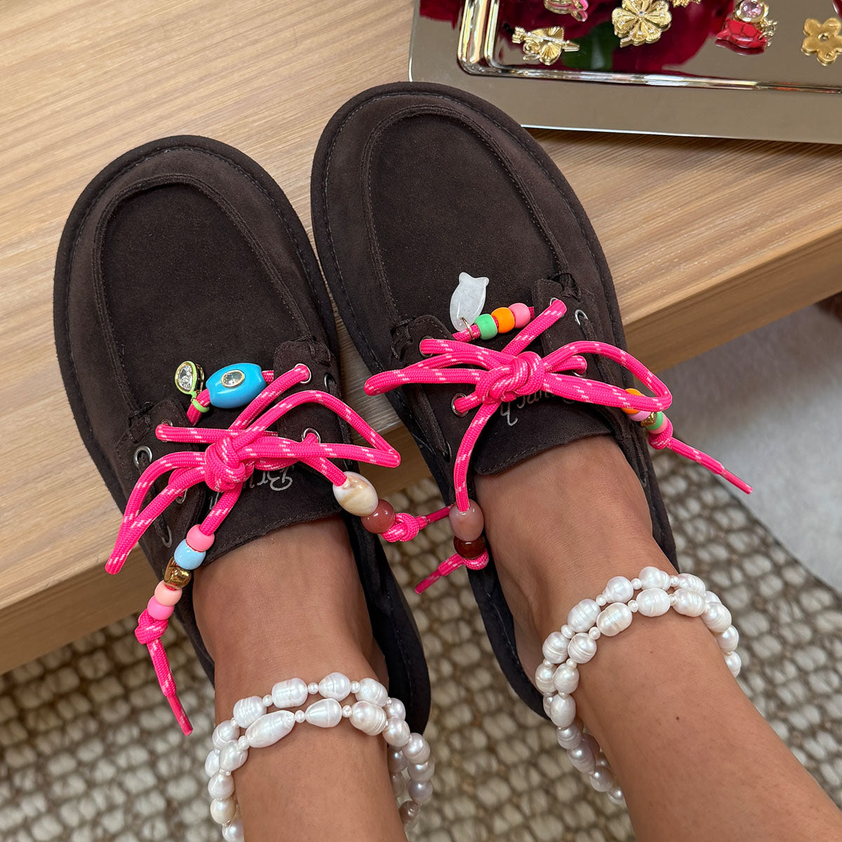 Top-down view of brown suede moccasin loafers showcasing neon pink beaded laces and pearl anklets on a wood surface